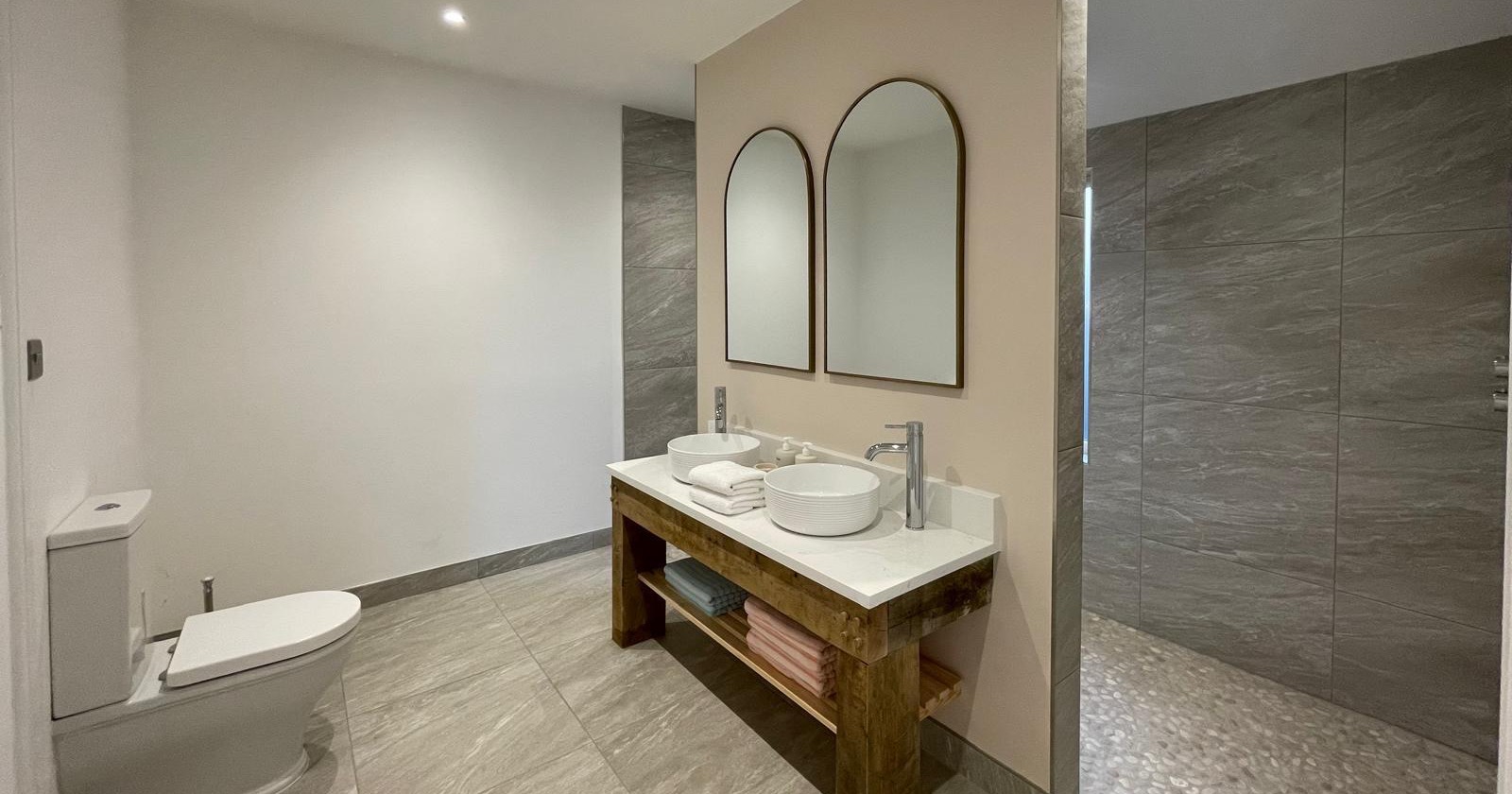 Double vanity with countertop basins on a rustic timber washstand, arched mirrors and a walk-in shower behind